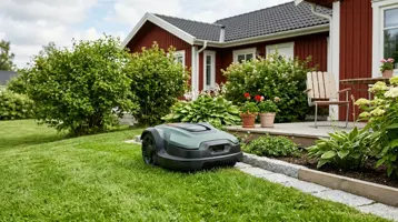 Robotgräsklippare på gräsmattan framför ett rött trähus med rabatter.