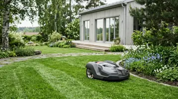 Robotgräsklippare på en randigt klippt gräsmatta framför ett modernt trähus.