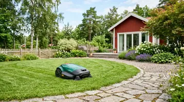 Robotgräsklippare på gräsmattan vid en stengång framför ett rött hus.