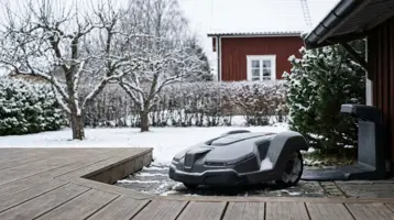 Robotgräsklippare uppställd på ett trädäck framför en snötäckt trädgård.